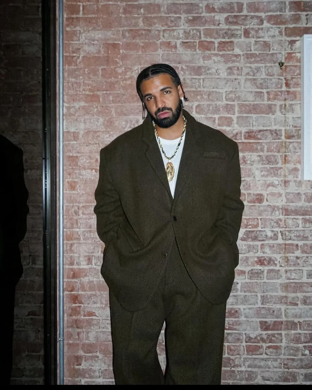 Man in oversized dark suit with braided hair standing against a brick wall.