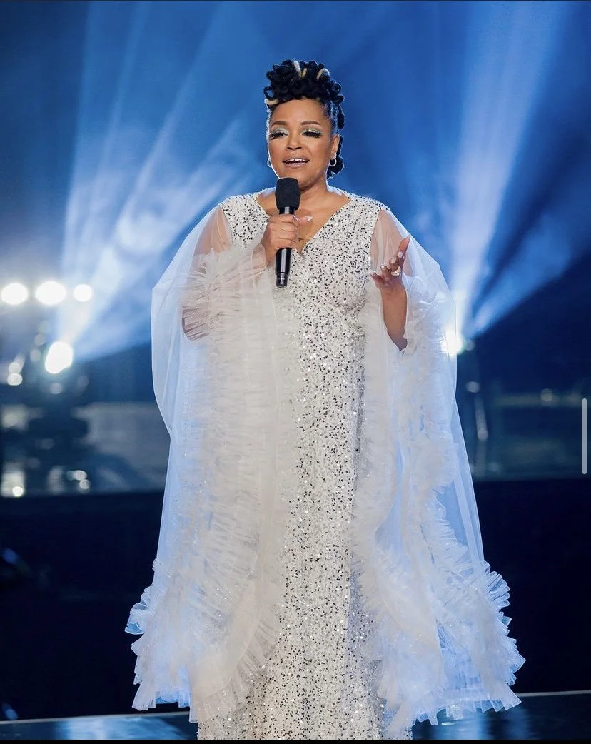 A person wearing a sparkly, embellished gown, holding a microphone, and standing on a stage with dramatic lighting and spotlights in the background.