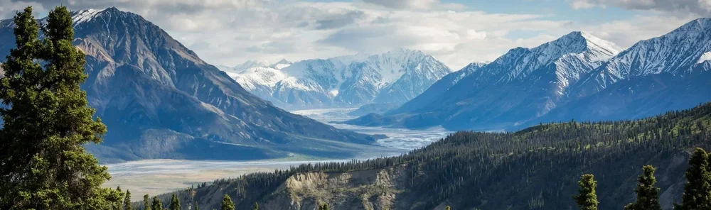 Snow-covered mountains with valley and evergreen trees in foreground.