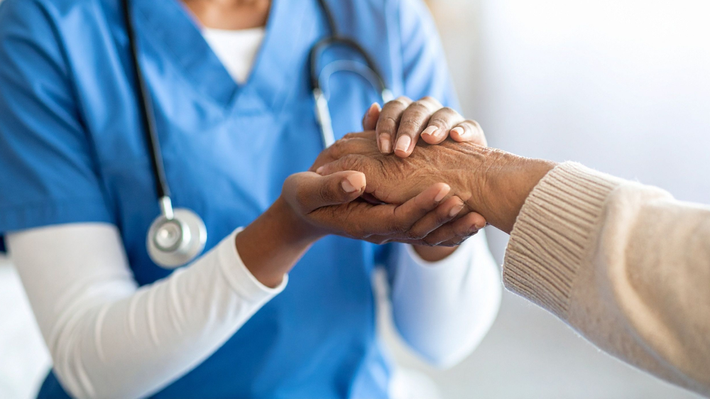 A healthcare professional in blue scrubs and a stethoscope holding an elderly person's hand, providing comfort or care.