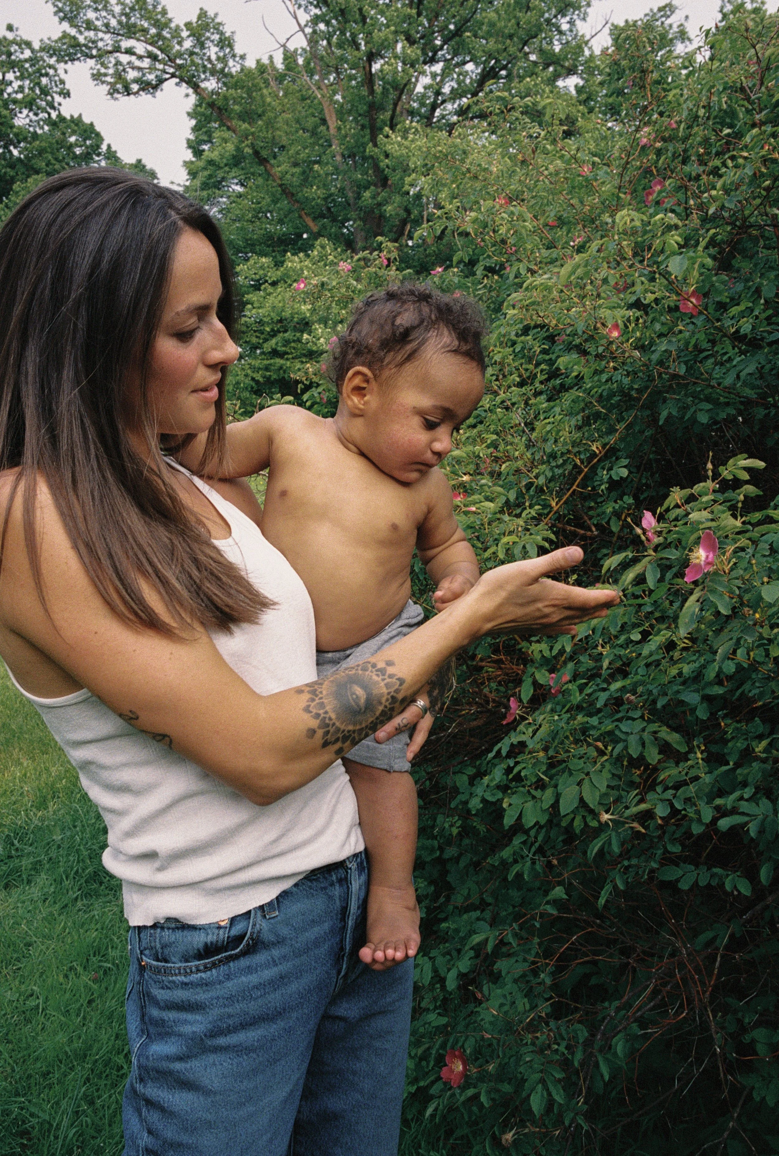 A woman with long brown hair, tattoos on her arm, and wearing a white tank top and blue jeans, holding a young child with curly hair and bare chest, in front of a bush with pink flowers and green trees.