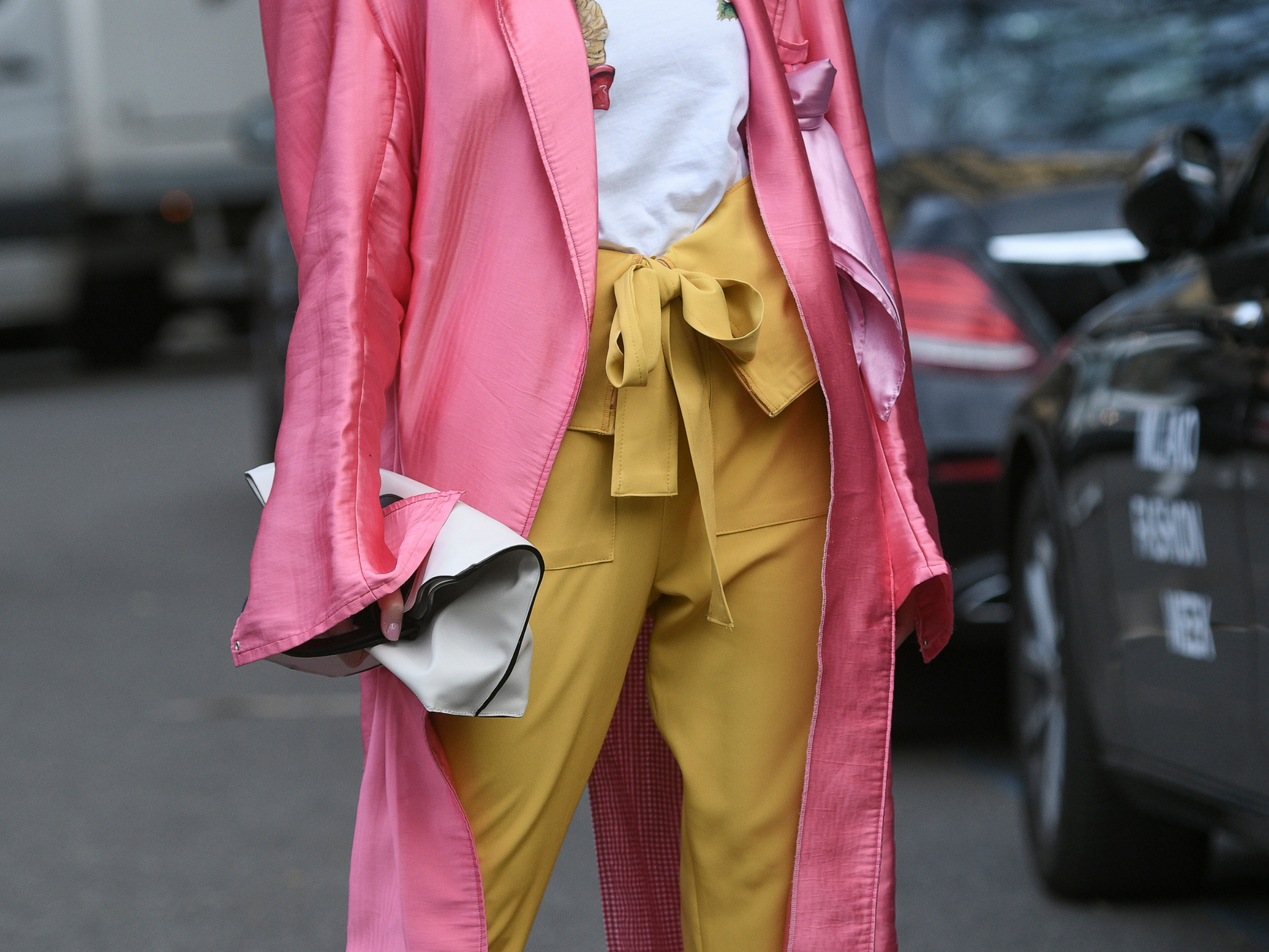 Person wearing a pink long coat, yellow pants with a matching yellow bow tied at the waist, and holding a black and white clutch bag, standing outdoors near parked cars.