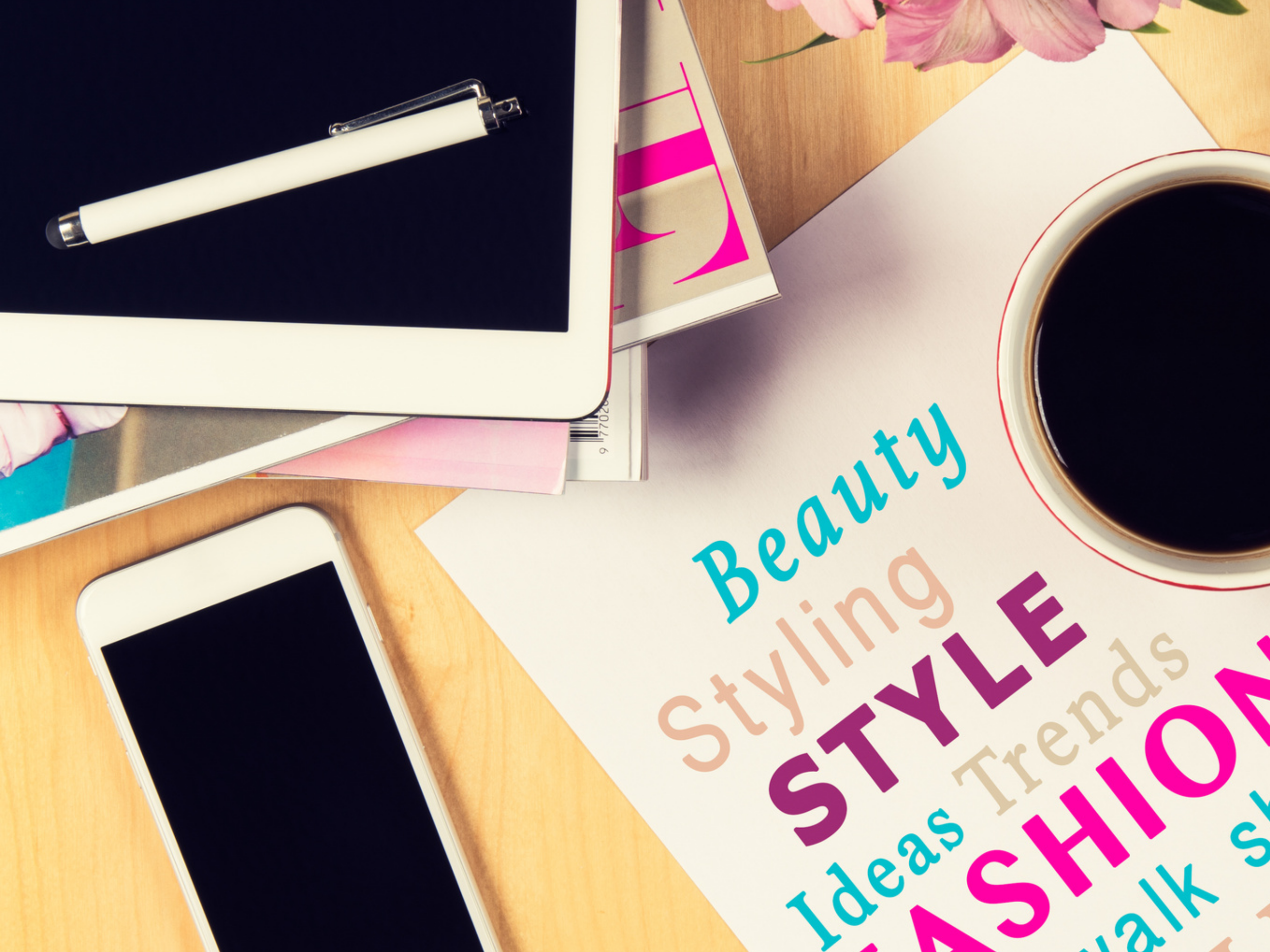Desk with a smartphone, tablet with stylus, magazines, a notepad with colorful words about beauty, styling, trends, and a cup of coffee.