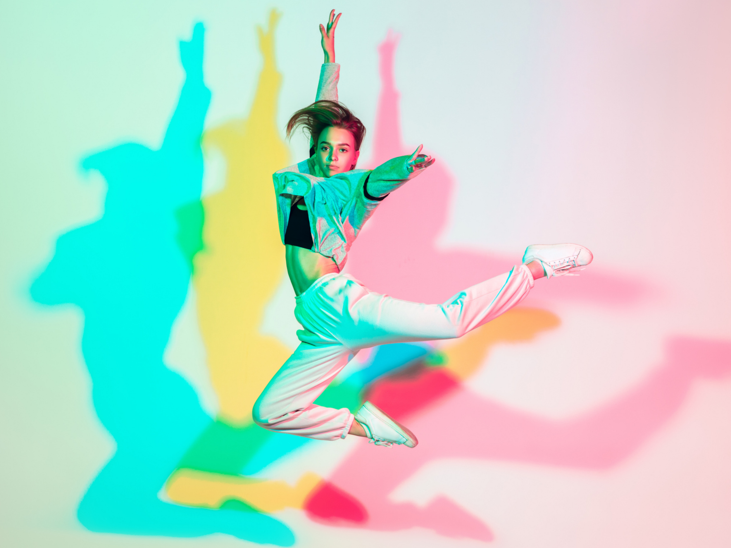 A young woman jumps in mid-air with her arms raised, wearing a colorful jacket, white pants, and sneakers, against a background with colorful shadows cast on the wall.
