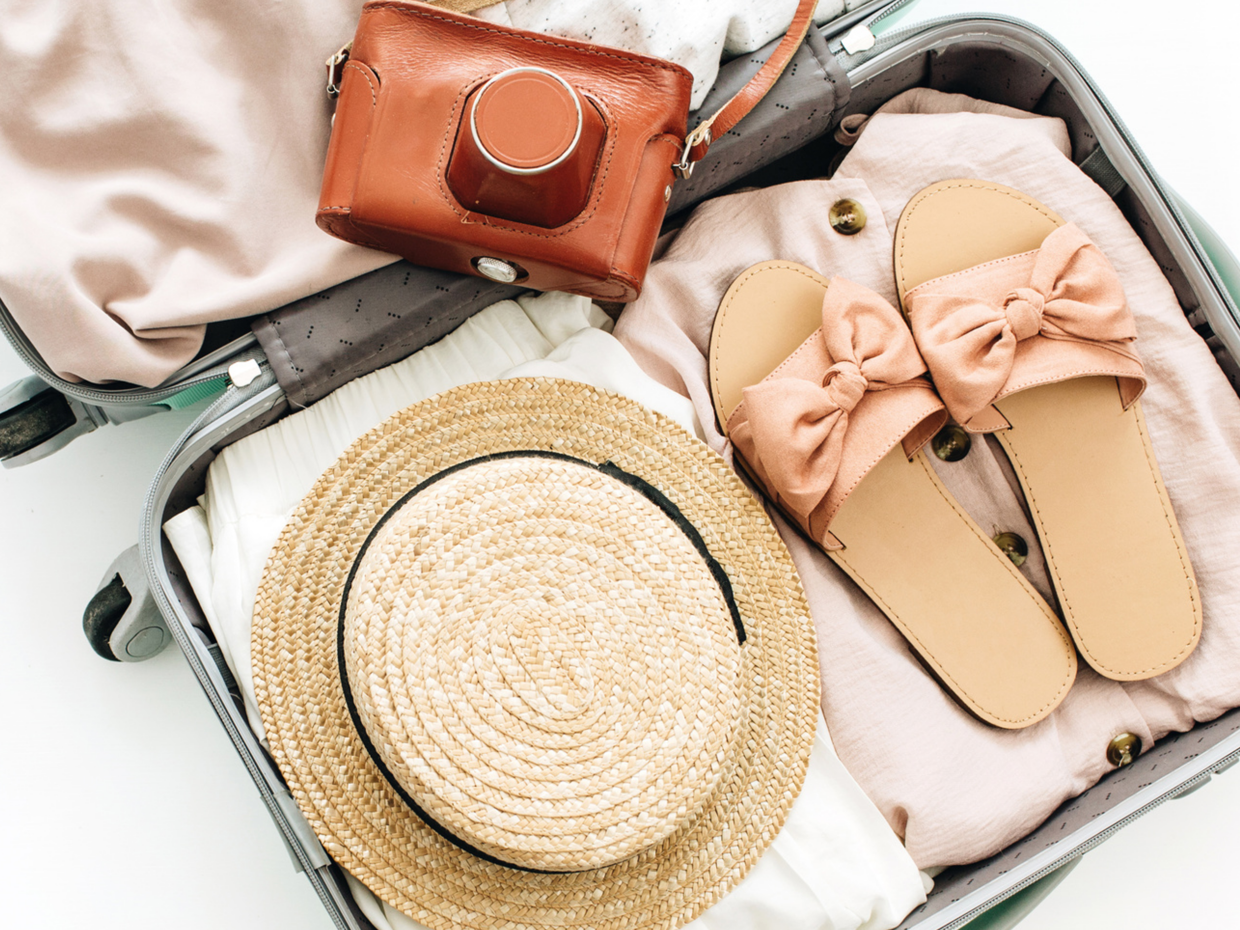 Open suitcase containing a straw hat, pink slides with bows, beige clothing, and a brown camera, indicating packing for a trip.