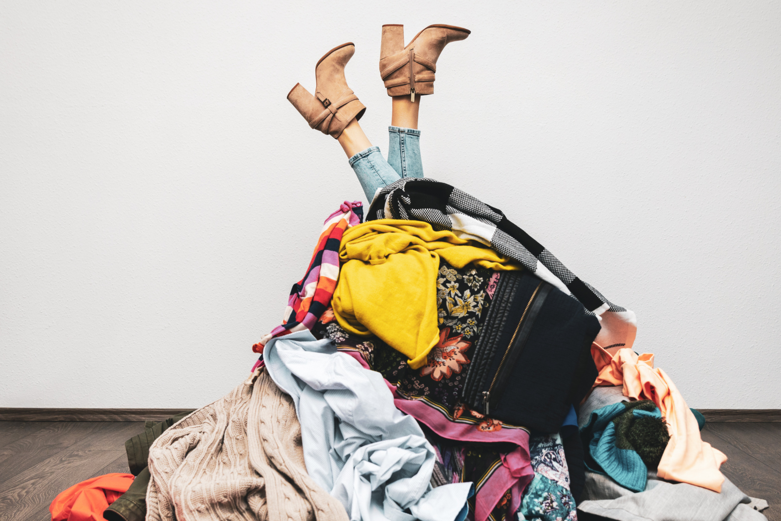A person's legs and feet in tan ankle boots with heels, sticking out from a pile of assorted clothes and fabrics on the floor, with the person lying underneath or amidst the clothes.