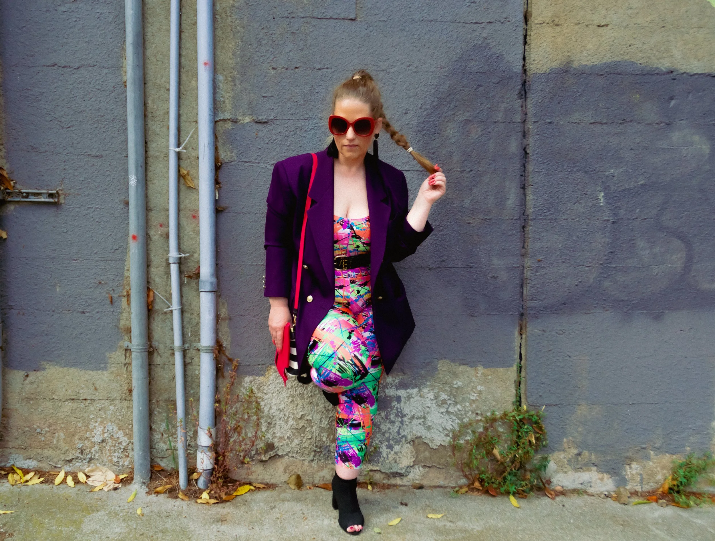 Woman dressed in a vibrant colorful dress and purple blazer, wearing large red sunglasses, standing against a rough gray wall with pipes and fallen leaves on the ground.