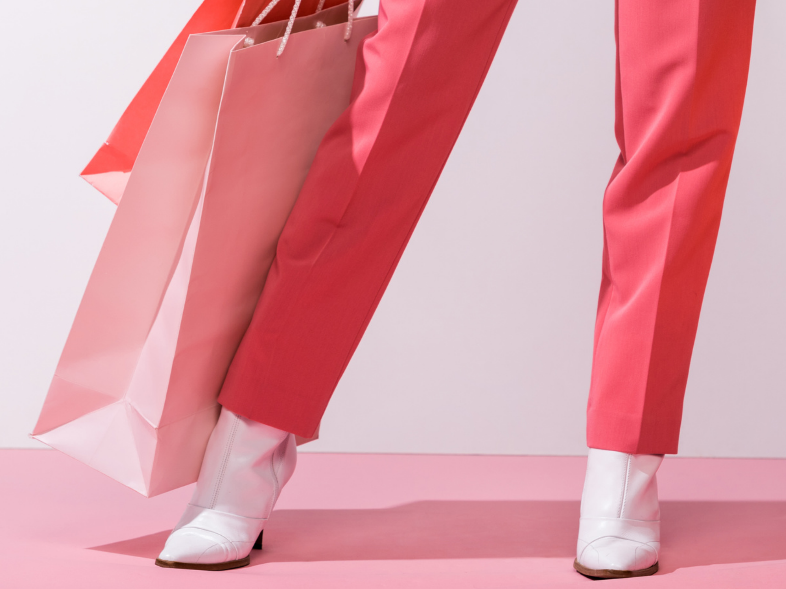 Close-up of a person wearing pink pants and white cowboy boots, holding pink shopping bags, standing against a pink and white background.