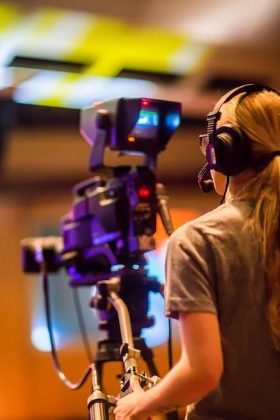 A woman operating a professional camera on a tripod, wearing headphones in a brightly lit indoor setting.