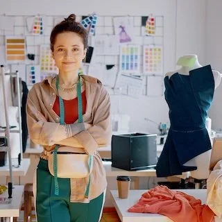 Young woman with brown hair and a green apron standing in a design workspace, smiling with arms crossed, with sewing patterns and fabric swatches on the wall behind her, and a mannequin dressed in a navy blue garment to her right.