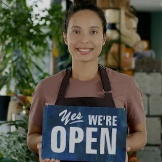 A smiling woman in a maroon shirt wearing an apron, holding a blue 'Yes We're Open' sign inside a shop with plants and shelves in the background.