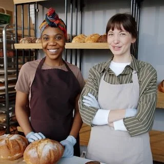 Two women standing behind a bakery counter with bread loaves in the background. One woman is African American with a colorful headscarf, wearing a brown apron, and smiling. The other woman is Caucasian with dark hair and bangs, wearing a striped green shirt and a light-colored apron, also smiling with arms crossed.