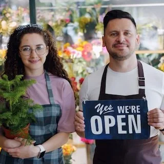 A man and a woman standing in a flower shop, holding a sign that says 'Yes, We're Open'. The woman is wearing glasses and a tiara, and the man is smiling. Both are wearing aprons.