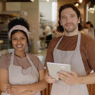 Two restaurant employees, a smiling woman and a man holding a tablet, standing behind the counter in a busy restaurant kitchen.