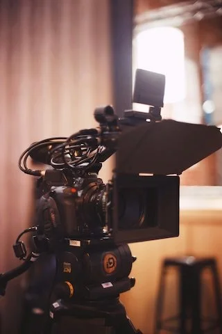 A professional video camera mounted on a tripod, set up in a room with a blurred background and natural light coming from a window.