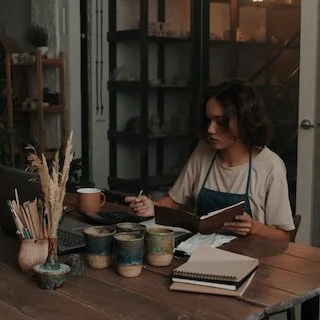 Woman at a wooden table writing in a notebook surrounded by stationery and craft supplies in a cozy workspace.