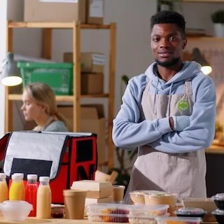 A man in a gray hoodie and apron standing in a kitchen or food prep area, with a woman working in the background.