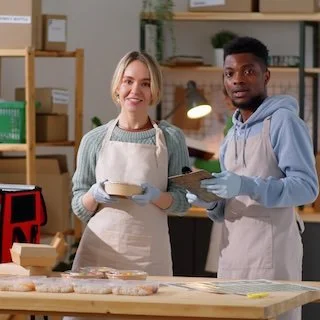 Two people wearing aprons and gloves in a kitchen or bakery, holding a tray and smiling at the camera.