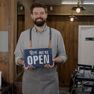 Man with a beard and light shirt holding a 'We're Open' sign inside a restaurant or cafe