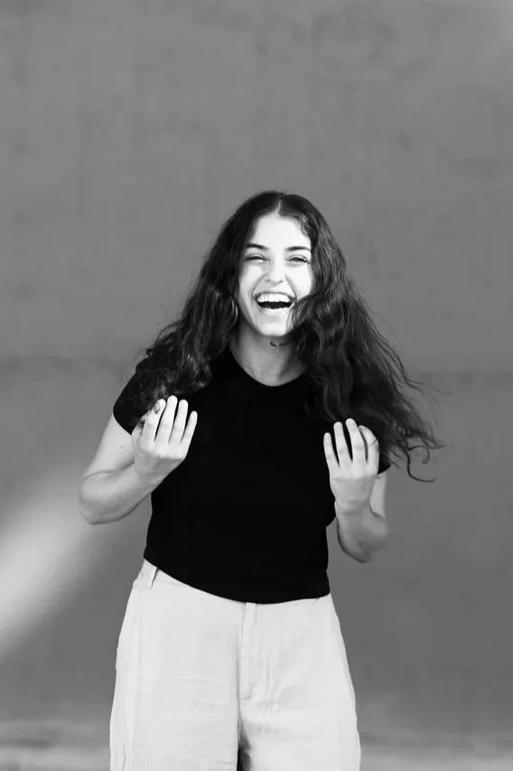 A woman with long, curly hair laughing and smiling, wearing a black t-shirt and light-colored pants against a plain background.