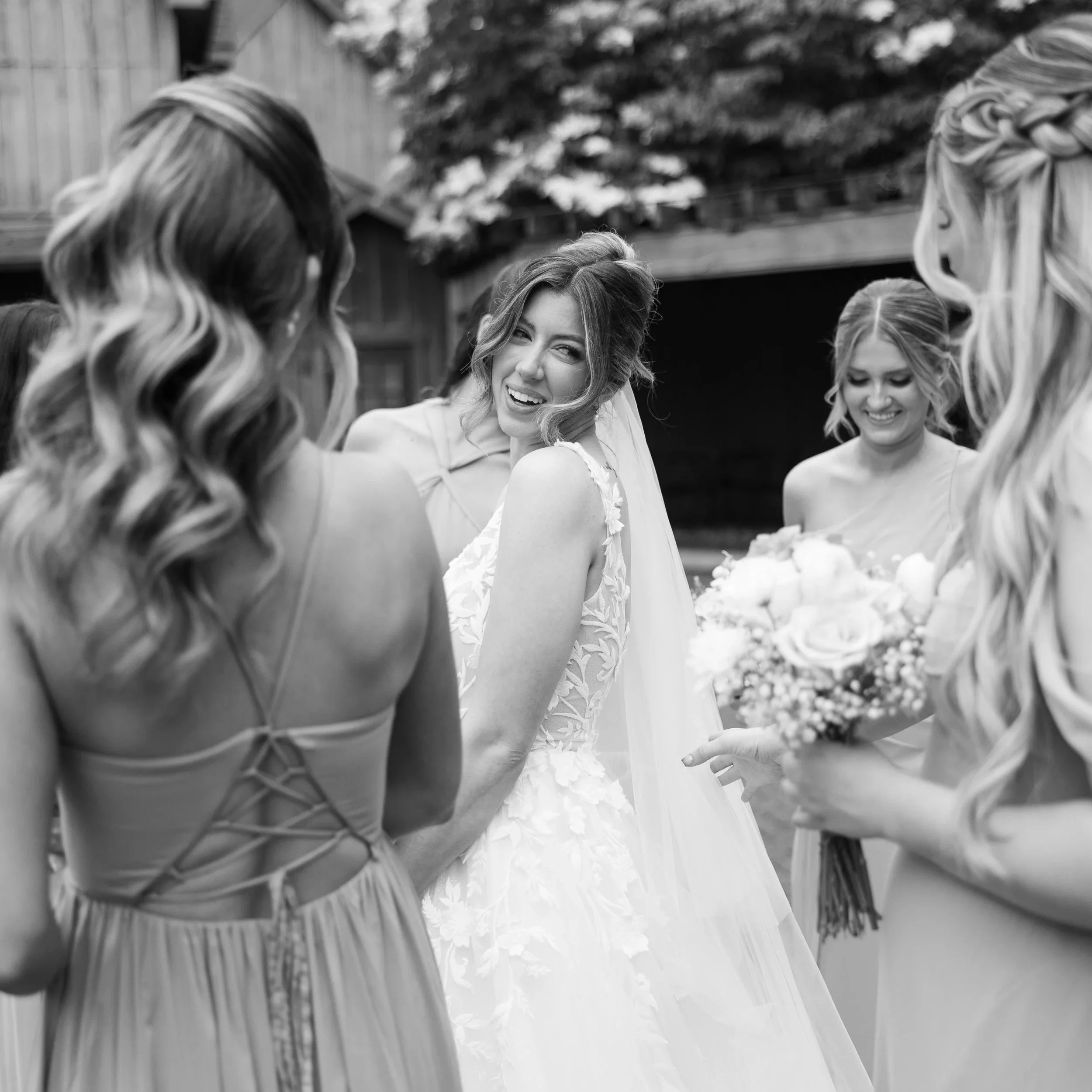 A bride smiling and winking at her bridesmaids during a wedding preparation outdoors.