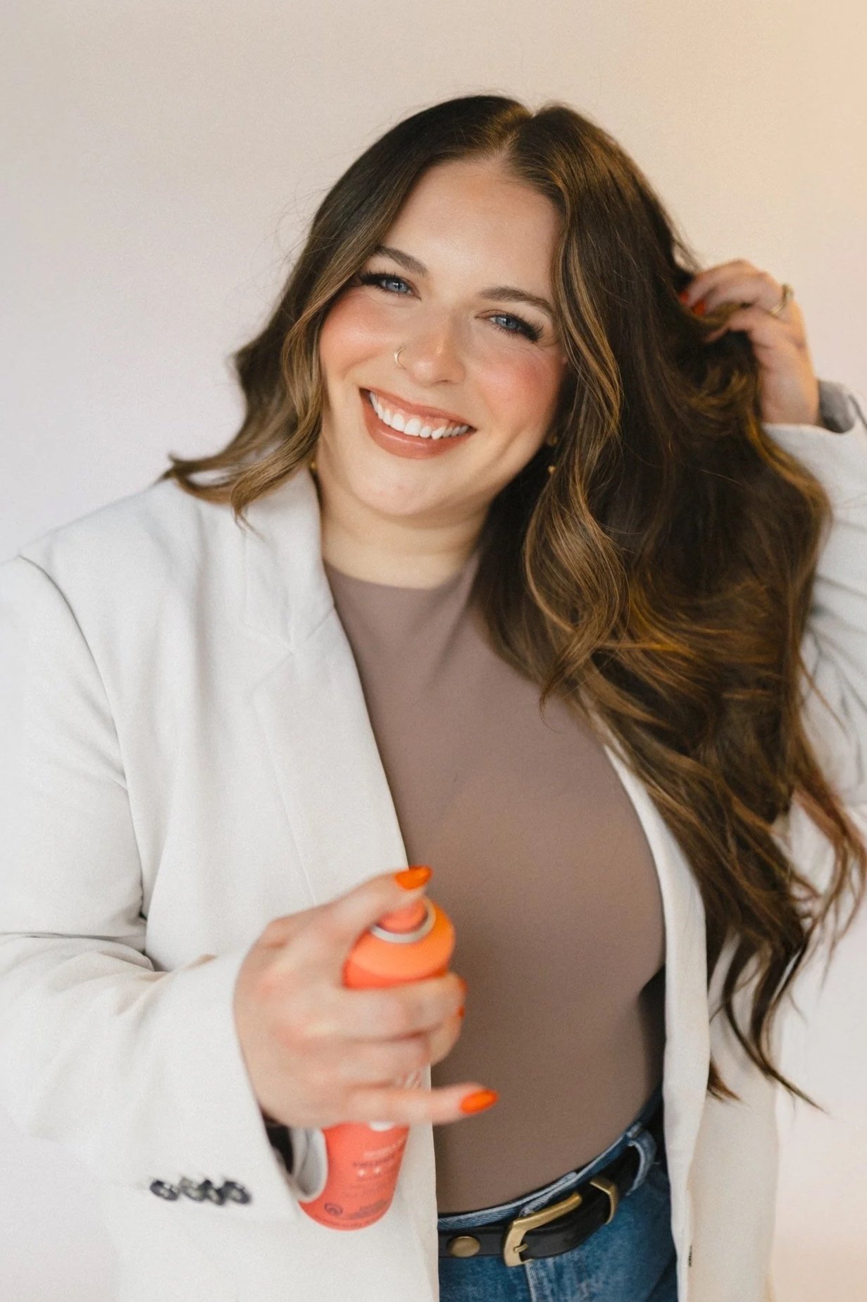 Smiling woman with wavy brown hair and blue eyes, wearing a white blazer over a brown top, holding an orange spray bottle, standing against a plain light background.
