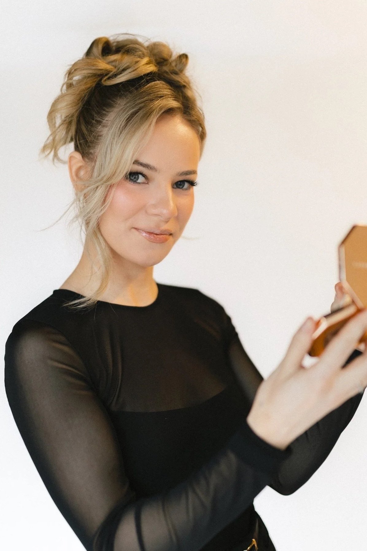A woman with blonde hair styled in an updo, wearing a black sheer long-sleeve top, holding a compact mirror, and looking at the camera with a slight smile.