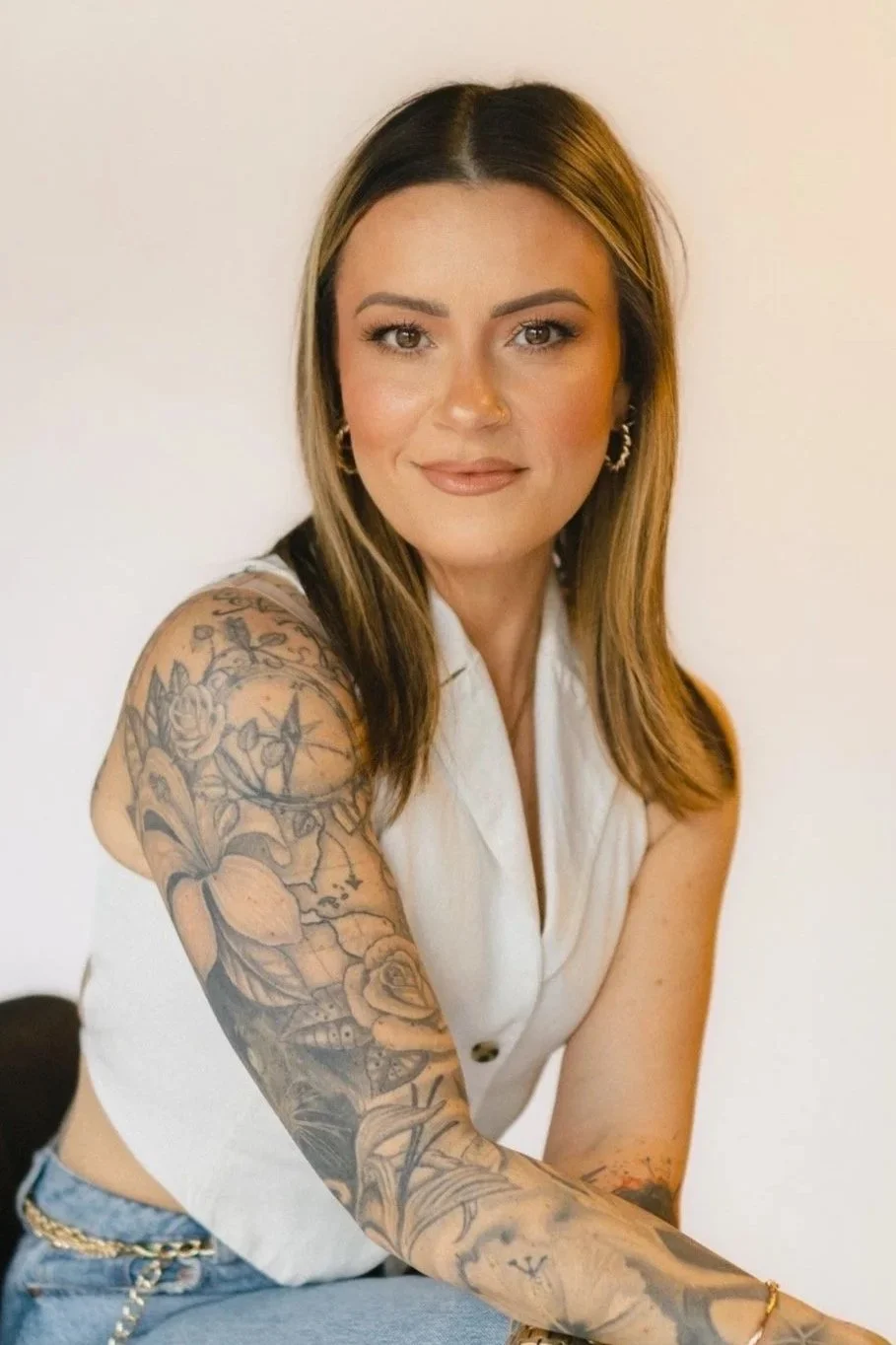 A woman with shoulder-length hair, wearing a sleeveless white top and light blue jeans, sitting against a plain white wall, with detailed tattoos on her arm, wearing jewelry and a watch.