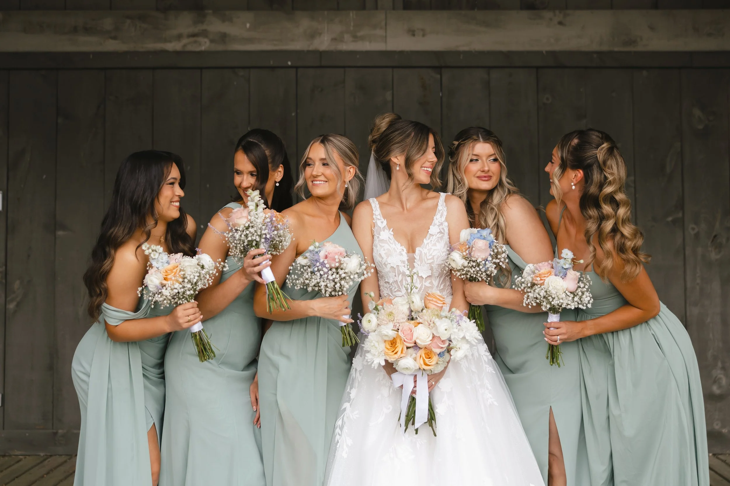 A bride with light brown hair in an updo and her five bridesmaids in light green dresses, standing together indoors against a dark wooden background, holding bouquets of flowers, smiling, and interacting happily.