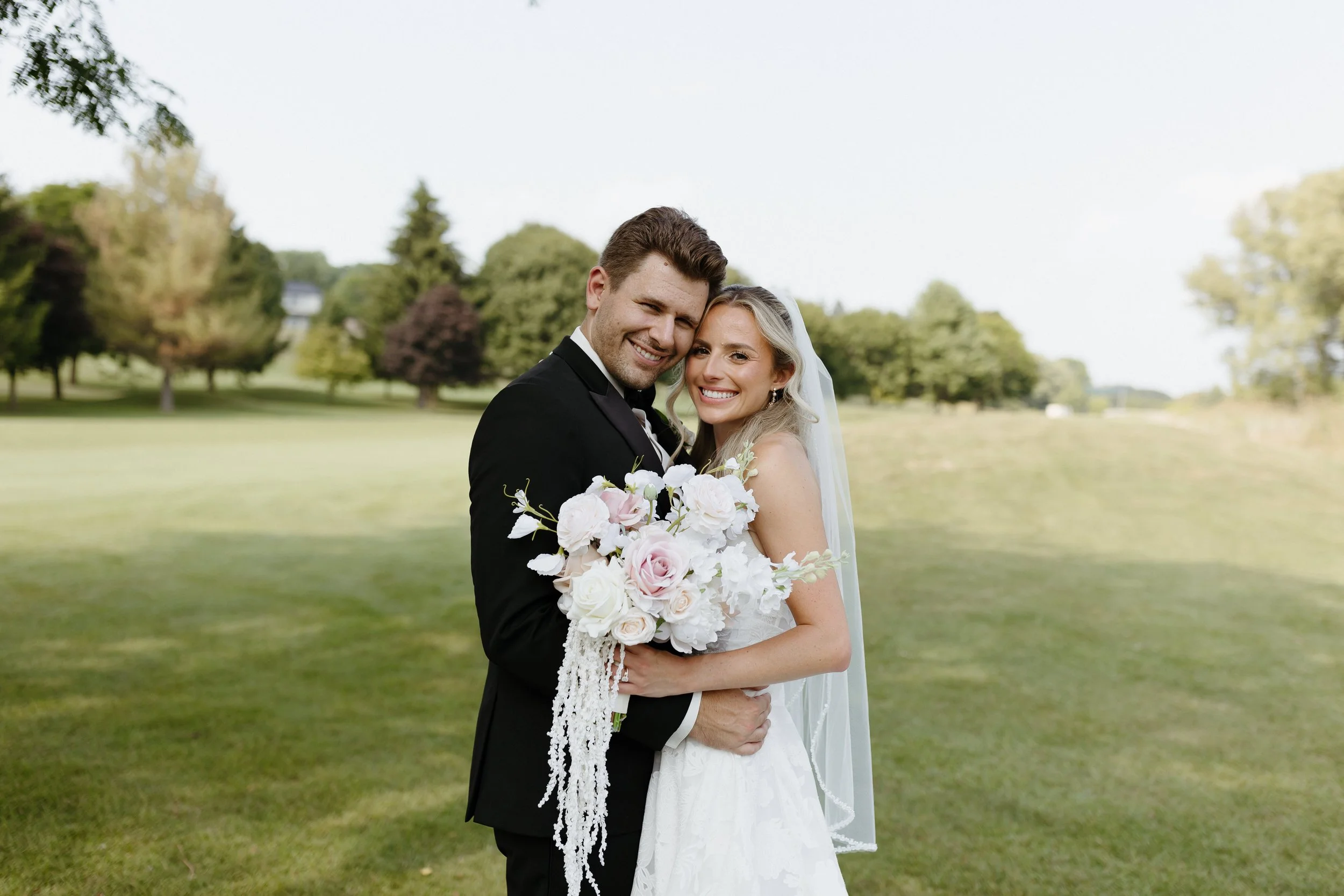 A newlywed couple smiling and embracing outdoors on a bright day, with the bride holding a large bouquet of white and pink flowers, standing on a grassy field with trees in the background.