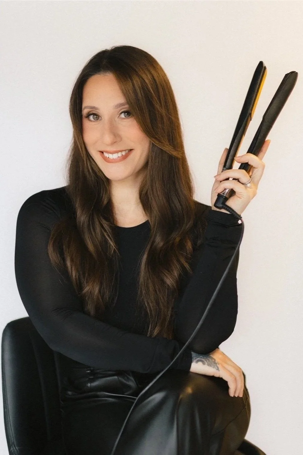 A woman with long brown hair, wearing a black top and leather pants, sitting on a black chair, smiling, holding a hair straightener.