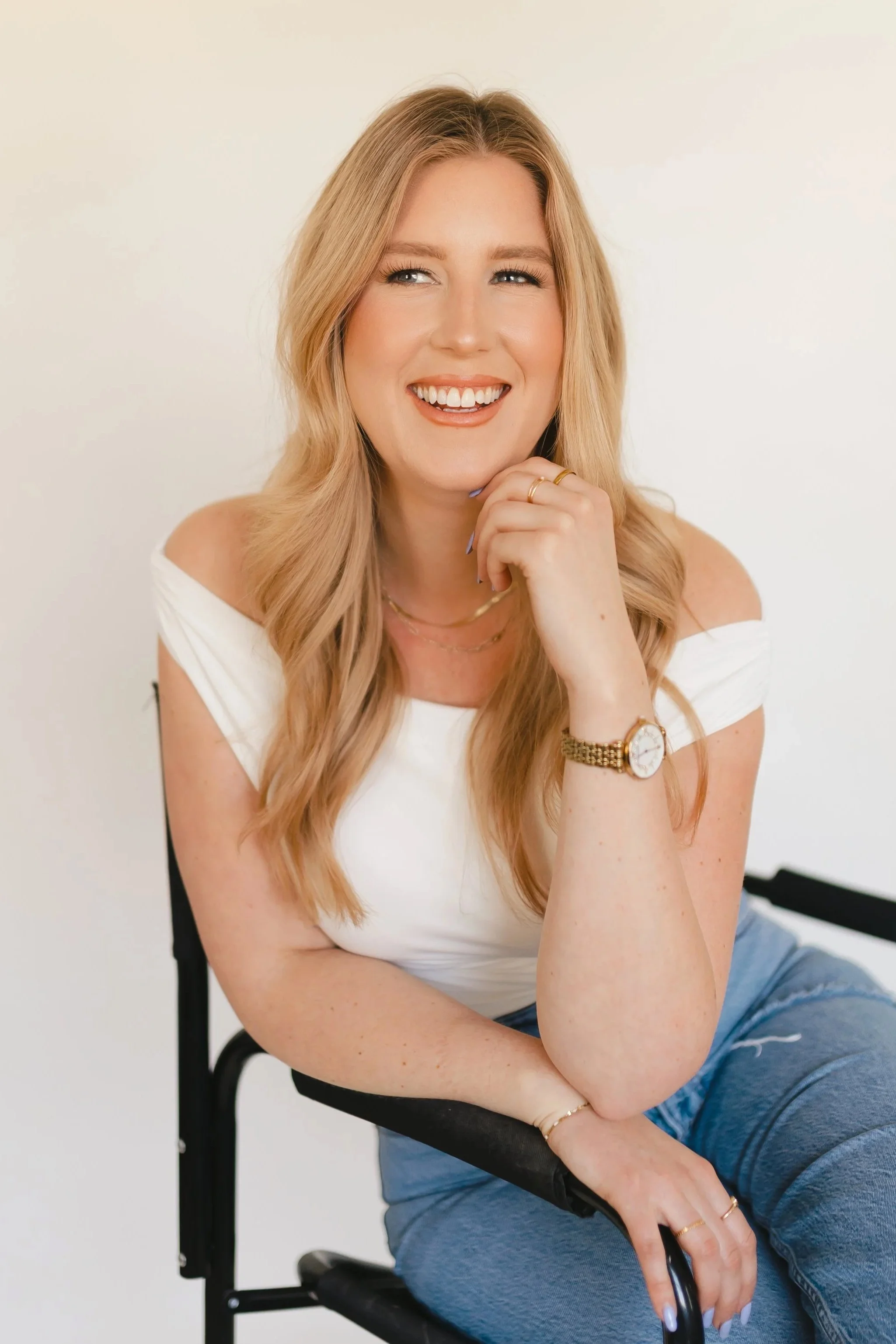 A woman with long blonde hair, wearing a white off-the-shoulder top and blue jeans, sitting on a black chair, smiling and looking to the side against a plain white background.