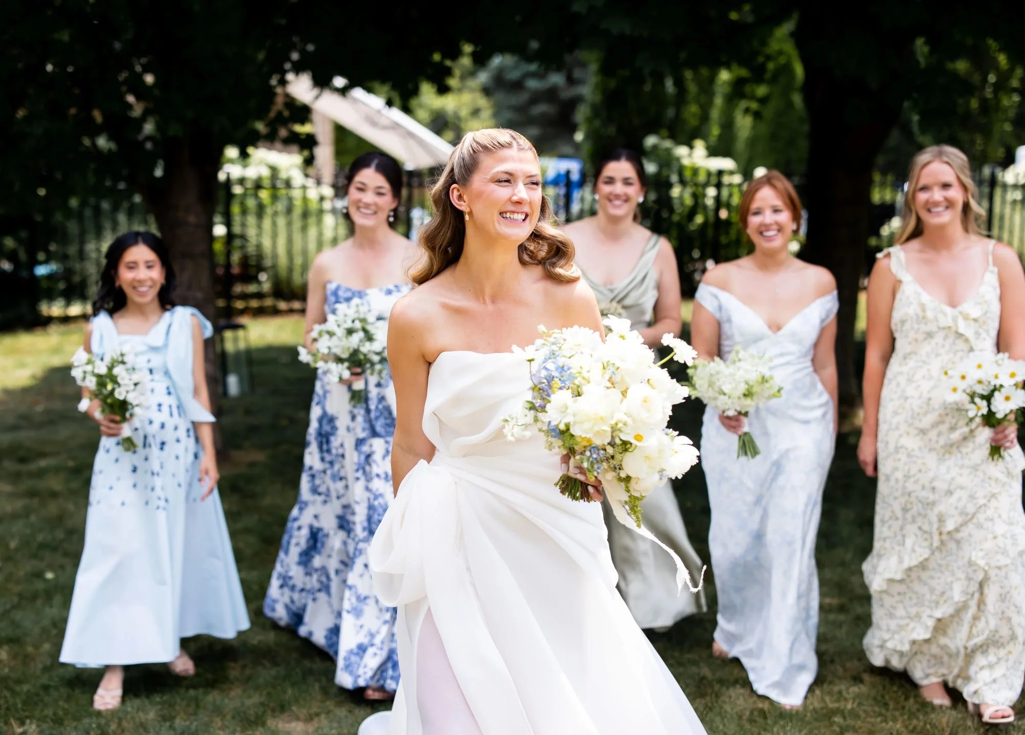 A bride in a strapless white wedding dress holding a bouquet of white and light purple flowers, walking outdoors on grass, with six women in colorful dresses behind her, all smiling and holding flowers, under a canopy of trees.