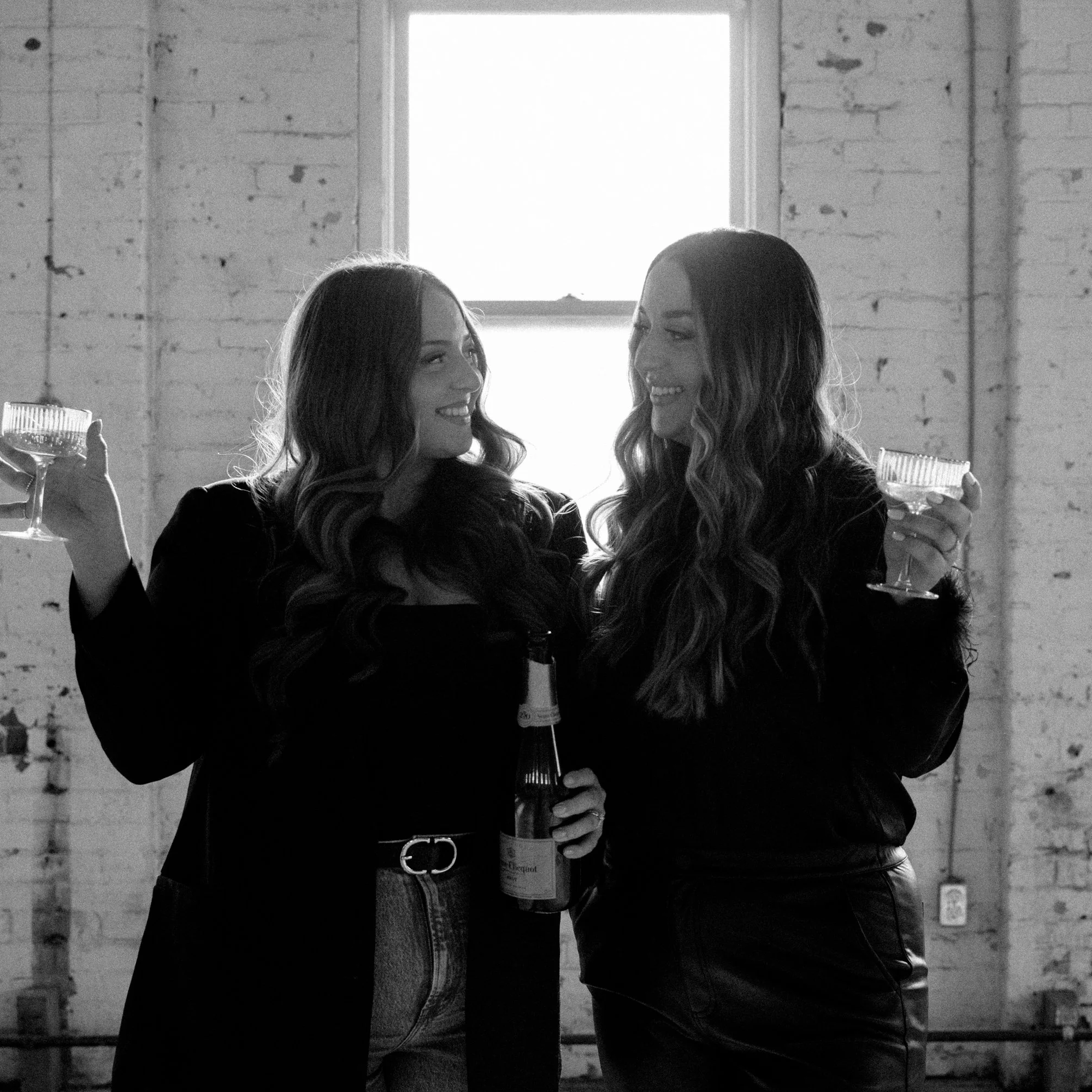 Two women smiling and holding champagne glasses in a room with a large window and brick walls, one woman holding a bottle of champagne.
