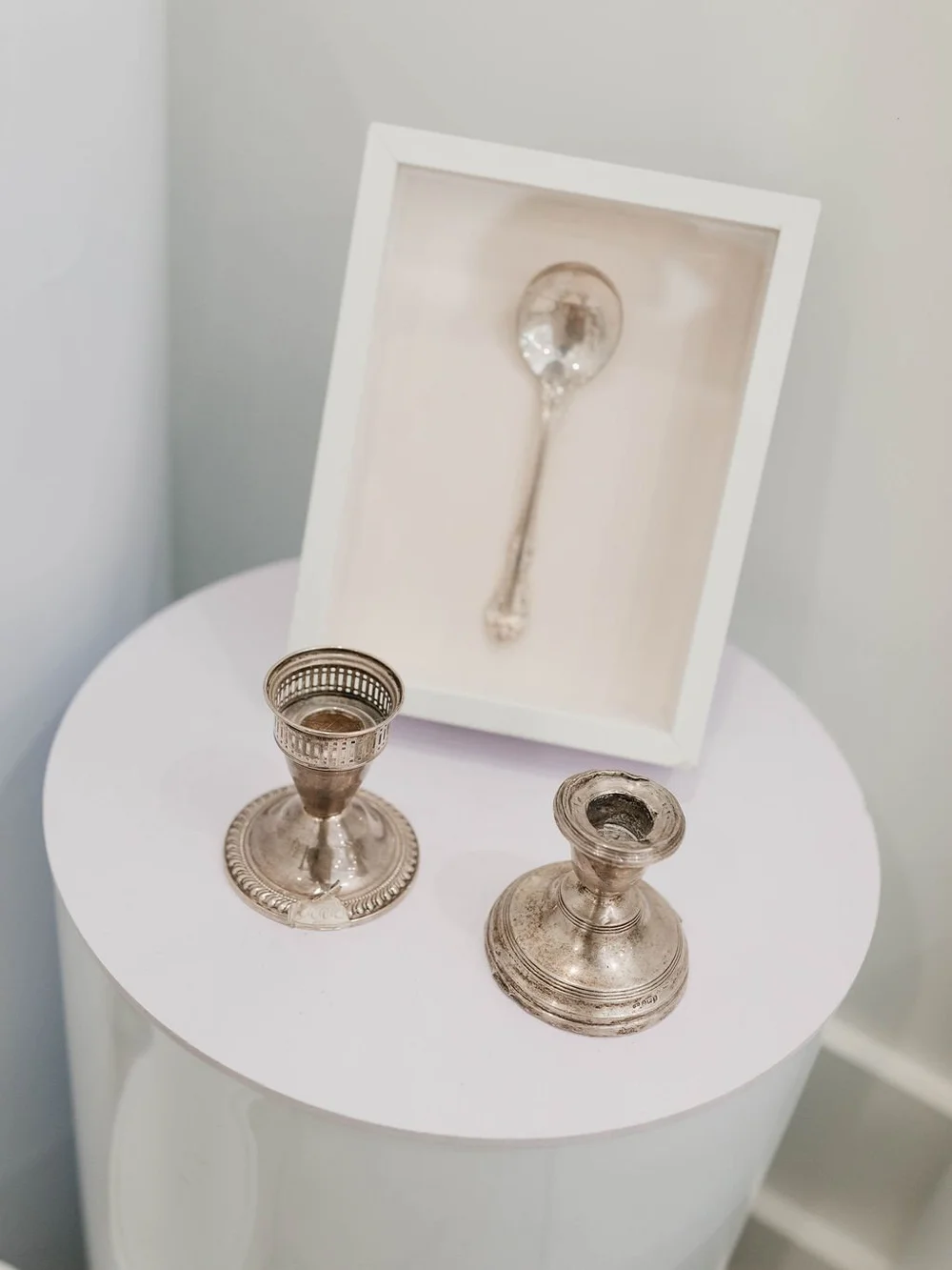 A silver spoon in a white shadow box on a round table beside two silver candlestick holders