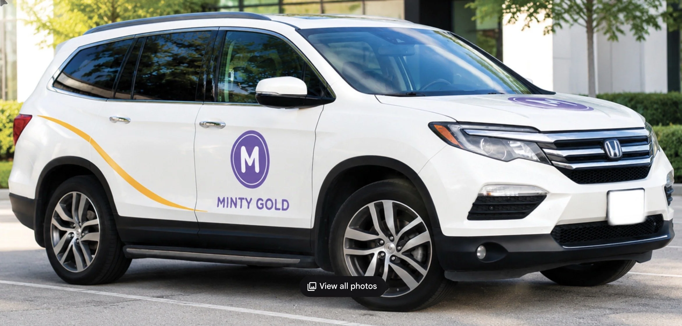 A white SUV with the logo of Minty Gold, a purple circle with an M in the center, parked outdoors on a paved lot.