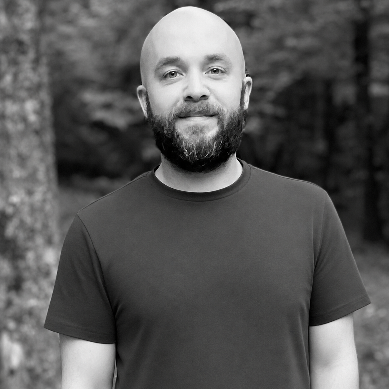 Black and white photo of Michael Hough with a beard, wearing a short-sleeved T-shirt, standing outdoors with blurred trees in the background.