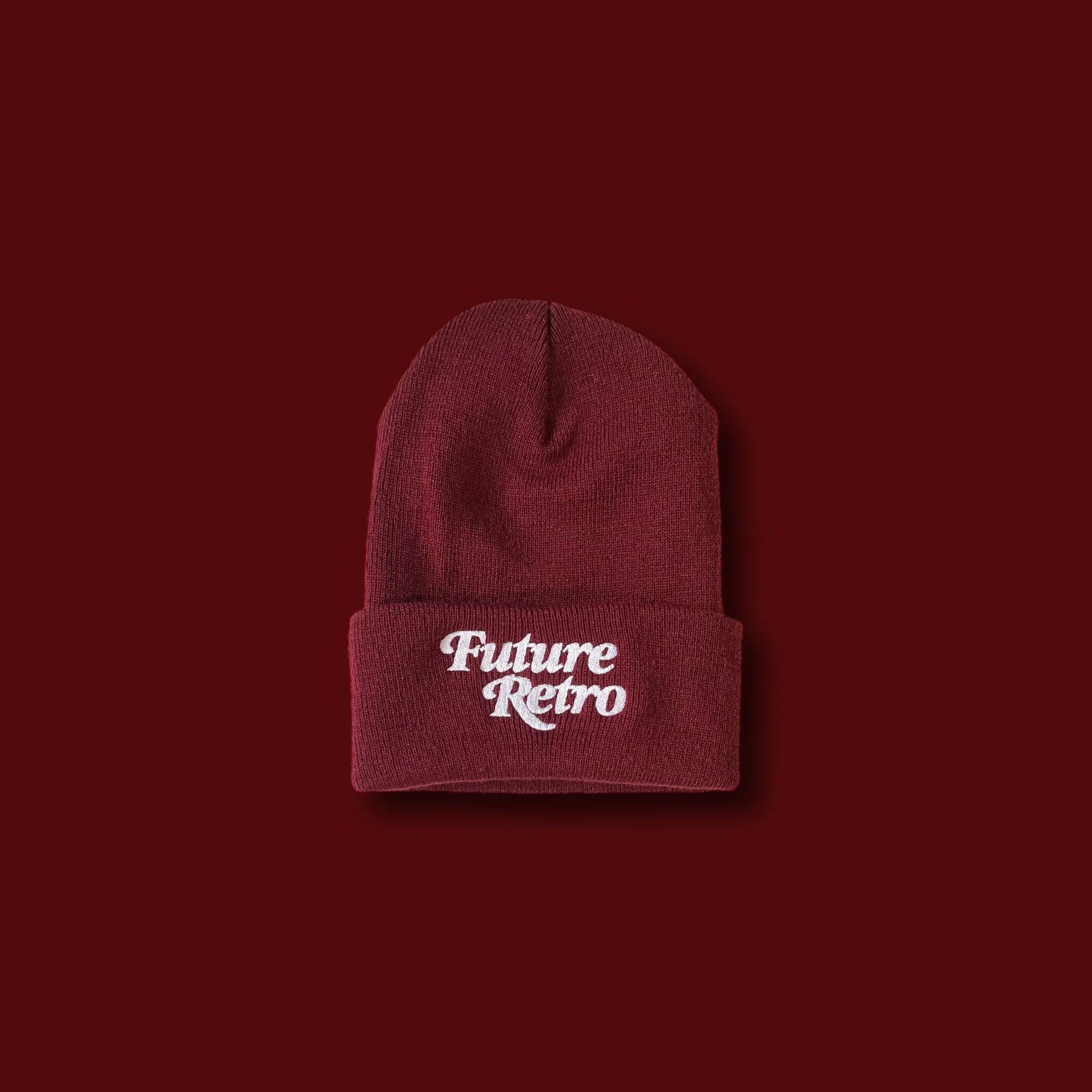 A maroon knit beanie hat with the words 'Future Retro' embroidered in white on the front centered cuff, placed on a matching maroon background.