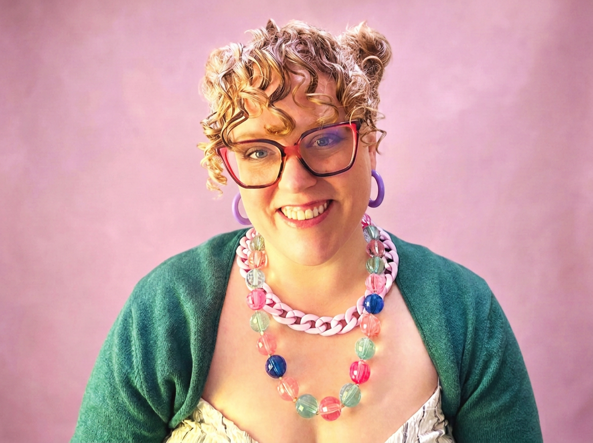 Coach Katrina smiles at the camera. She's a cis woman with greying blonde curly hair in space buns, with dark blue eyes. She wears a teal sweater over a striped sundress with an oversized double strand pink & blue bead & chain necklace, and glasses.