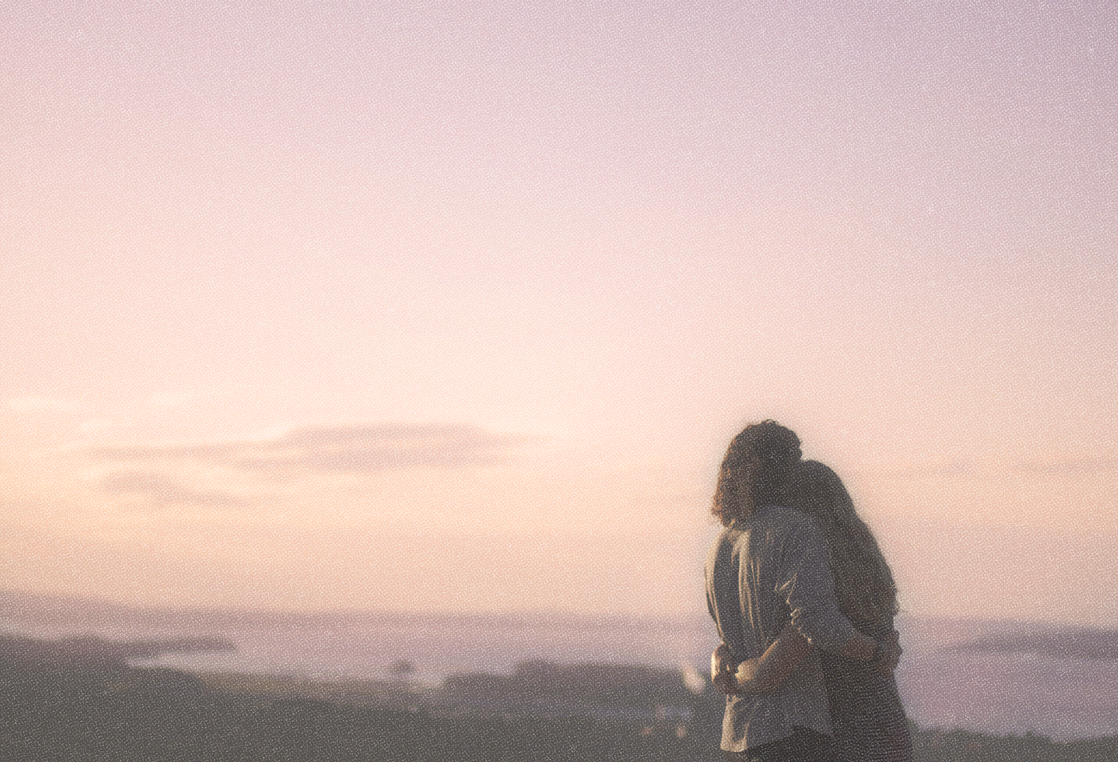 Image of a couple hugging and looking out from above at a body of water and a pink sky at sunset