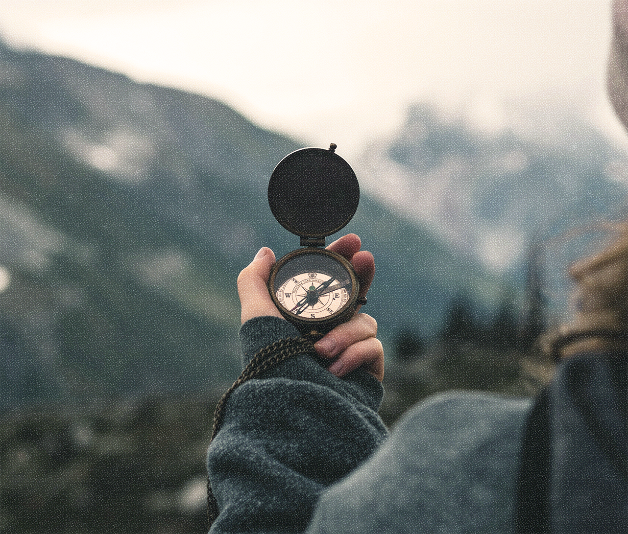 An image of a woman's hand holding a compass with mountains and trees blurred in the background