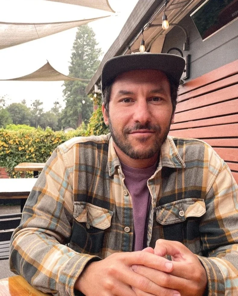 A seated portrait of Dustin Adler wearing a brown flannel shirt and a hat.