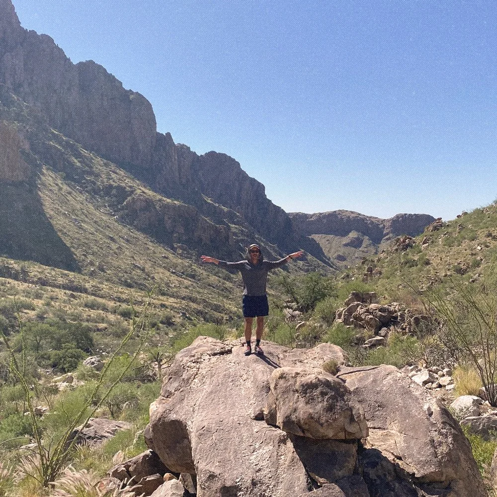A man hiking in a mountainous desert stands atop a large boulder with arms joyfully outstretched.