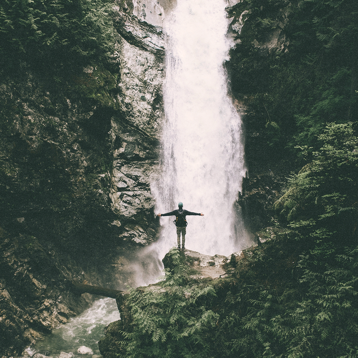 A hiker stands joyfully with arms outstretched, facing a large waterfall on a rocky mountainside.