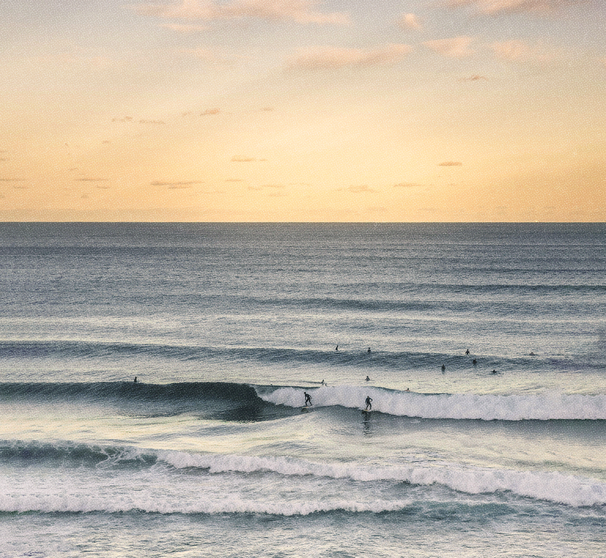 Sunset over the ocean with several surfers riding waves and others waiting in the water.