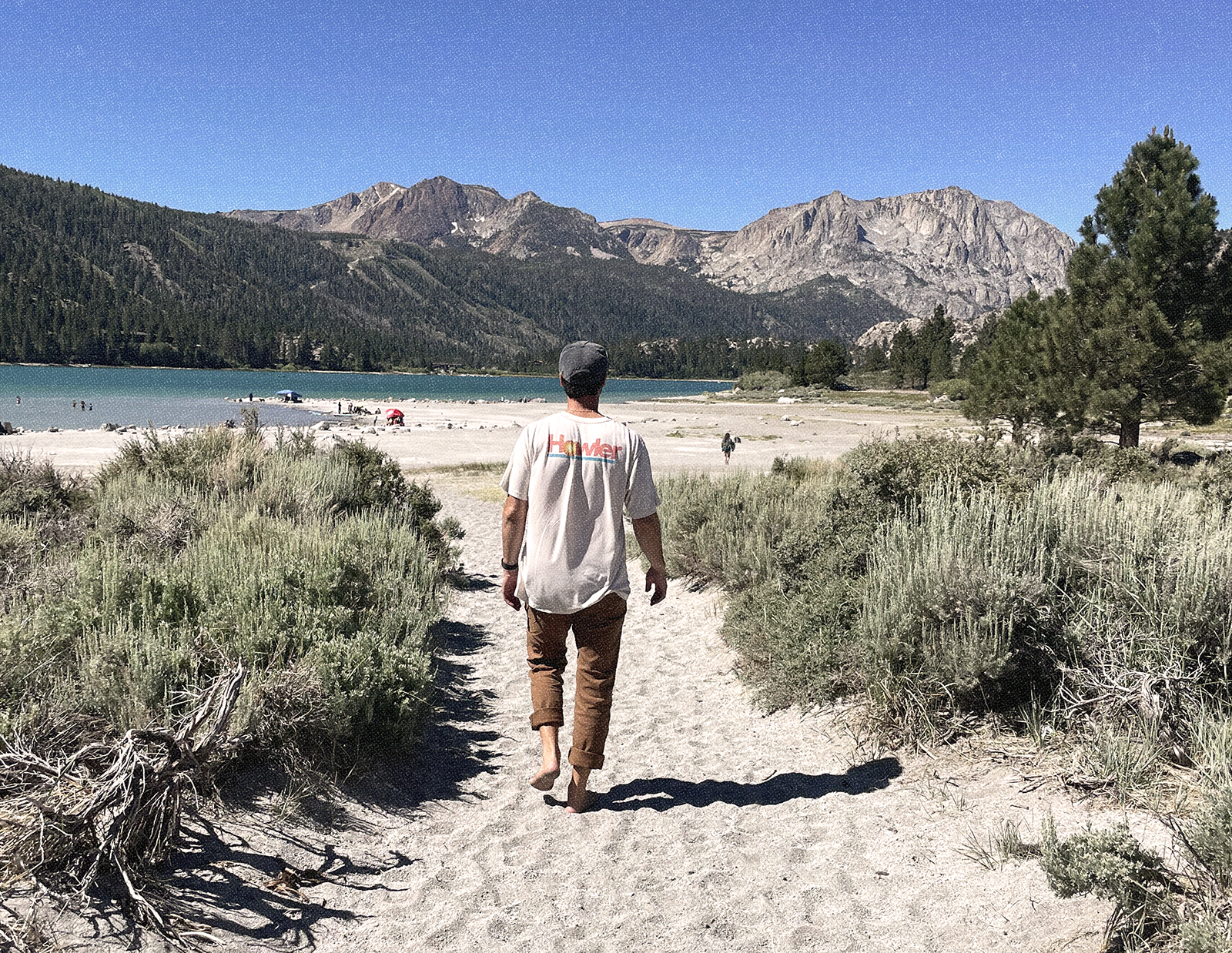 A barefoot man walking toward a lake on a sandy shore, surrounded by mountains.
