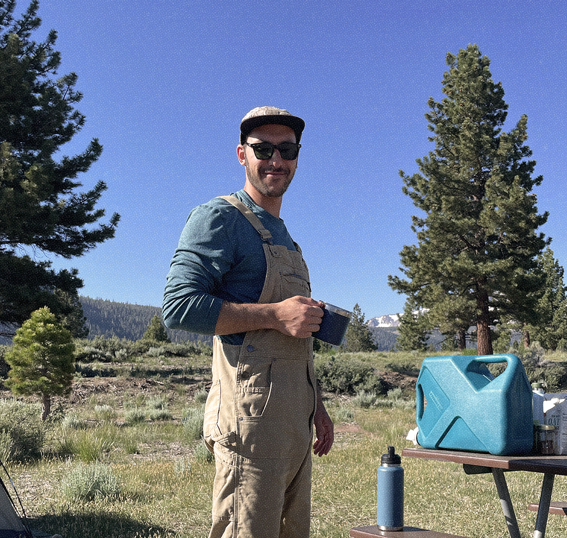 A man standing outdoors in a forested area with tall pine trees, holding a cup, wearing sunglasses, a baseball cap, a teal long-sleeve shirt, and tan overalls. There is a blue water container on a table next to him, with a smaller bottle nearby, and a mountain range visible in the distance under a clear blue sky.