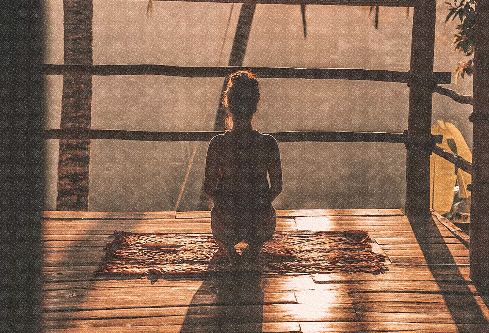 An image of a woman's silhouette resting in a meditative pose, overlooking a tropical landscape during golden hour.