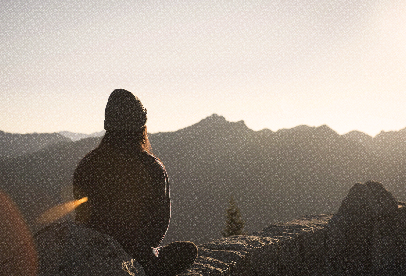 Image of a woman in a beanie sitting on a mountaintop looking out at the sunrise or sunset.