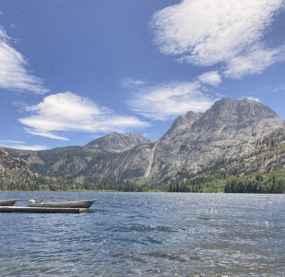 Image of a small silver boat floating in a lake with mountains and greenery in the background. There are soft white clouds against a blue sky.